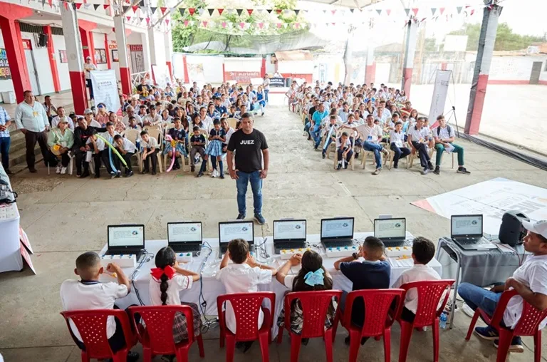 Un grupo numeroso de estudiantes y adultos sentados en un patio escolar cubierto observan una actividad educativa. En primer plano, varios niños están sentados frente a mesas con computadores portátiles, participando en una demostración. Al centro, un adulto de pie coordina la actividad. El espacio está decorado con banderines y se percibe un ambiente comunitario y escolar.