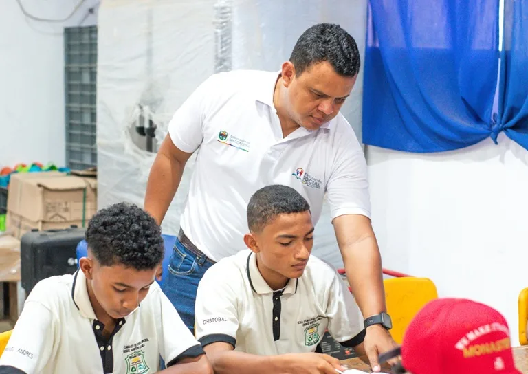 Dos estudiante en un salón de clases sentados, reciben explicación de un docente.